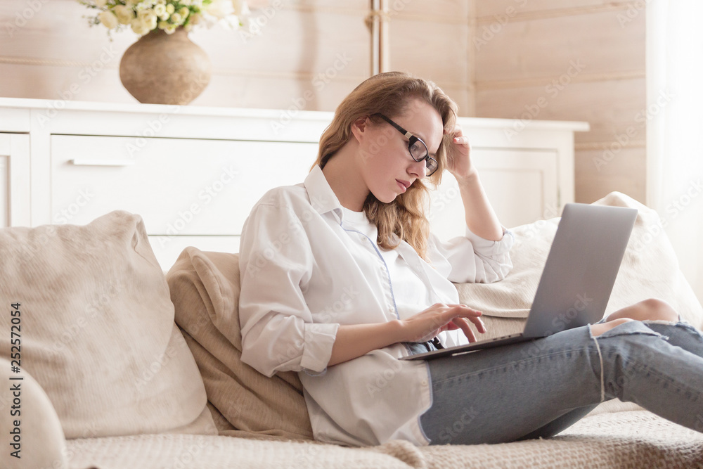 Pensive young woman freelancer working remotely using a laptop while sitting on a sofa in her cozy living room. Concept of comfortable work at home Pensive young woman freelancer working remotely using a laptop while sitting on a sofa in her cozy living room. Concept of comfortable work at home