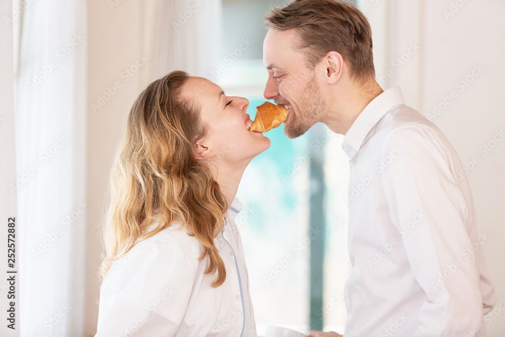 Side view of a funny happy married couple biting a croissant simultaneously from two sides. Concept of breakfast and good relationship Side view of a funny happy married couple biting a croissant simultaneously from two sides. Concept of breakfast and good relationship