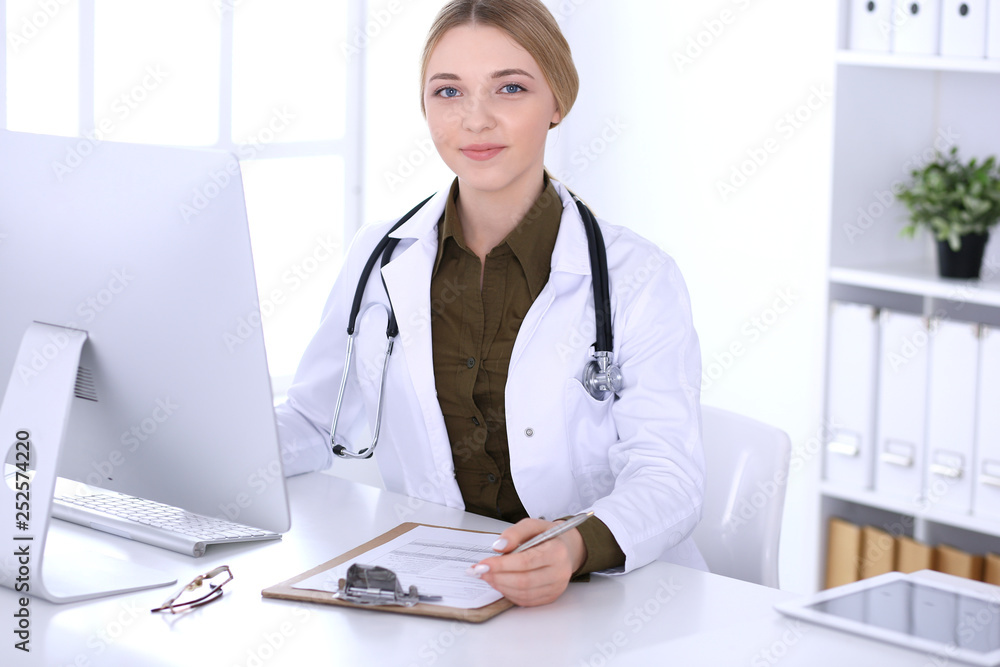Young woman doctor at work in hospital looking at desktop pc monitor. Physician controls medication history records and exam results. Medicine and healthcare concept Young woman doctor at work in hospital looking at desktop pc monitor. Physician controls medication history records and exam results. Medicine and healthcare concept