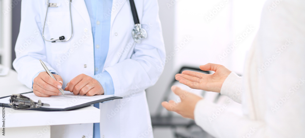 Unknown woman doctor and female patient discussing something while standing near reception desk in emergency hospital. Physician at work in clinic. Medicine and health care concept Unknown woman doctor and female patient discussing something while standing near reception desk in emergency hospital. Physician at work in clinic. Medicine and health care concept