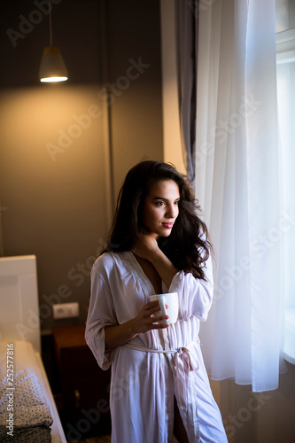 Young woman with mug by the window in bedroom