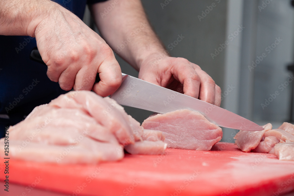 Closeup cook hand cuts pork fillet steak with sharp knife on red plastic cutting board on metal table in restaurant kitchen. Concept steakhouse specializing in grilled meat, live fire