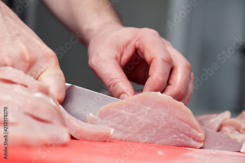 Closeup cook hand cuts pork fillet steak with sharp knife on red plastic cutting board on metal table in restaurant kitchen. Concept steakhouse specializing in grilled meat, live fire