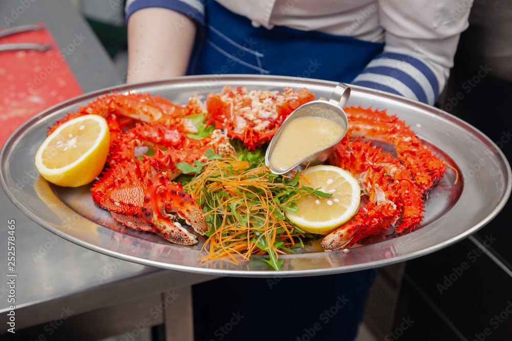 Chef cook in uniform hold metal dish with fresh red big boiled prepared blue kamchatka crab with greens, lemon. Concept useful seafood menu premium restaurant preparing, serving for guests, clients Chef cook in uniform hold metal dish with fresh red big boiled prepared blue kamchatka crab with greens, lemon. Concept useful seafood menu premium restaurant preparing, serving for guests, clients