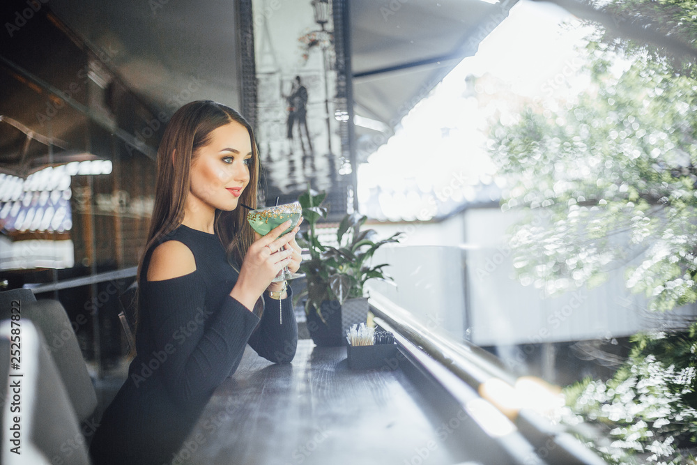 Young beautiful girl drinking a refreshing cocktail on a summer terrace cafe in a small black dress Young beautiful girl drinking a refreshing cocktail on a summer terrace cafe in a small black dress