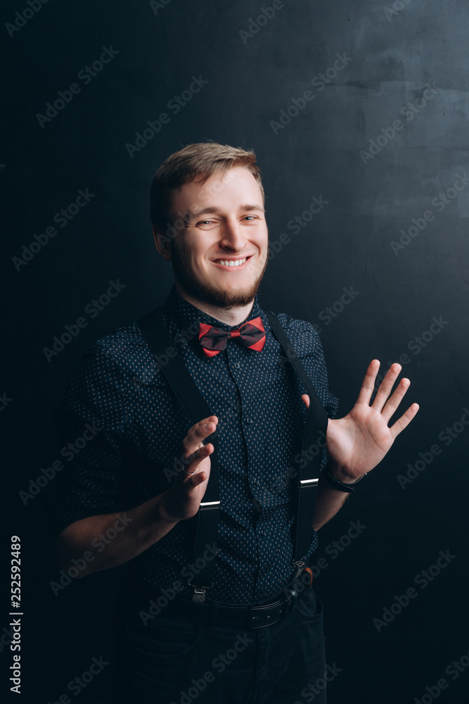 Portrait of young happy handsome stylish man smiling in dark blue shirt with suspenders and a butterfly tie on black background