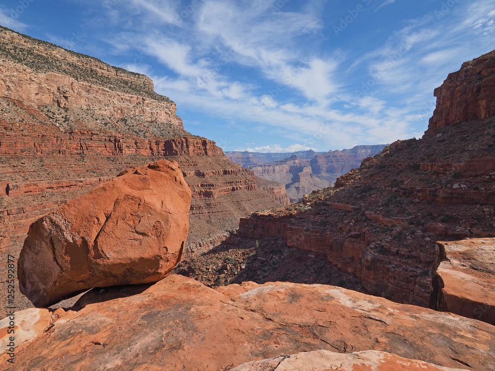 A large boulder perched on a cliff edge on the Hermit Trail in Grand Canyon National Park surrounded by red canyon walls under a brilliant cloudscape.