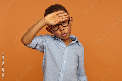 Human facial expressions and body language. Handsome African American male child in round eyeglasses having relieved look, breathing air out, making Phew sound, whiping sweat from his forehead