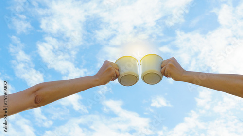 Two Asian woman hands clink hot coffee mug outdoor in the morning. Friends enjoy drinking coffee together. Clear sky background. photo