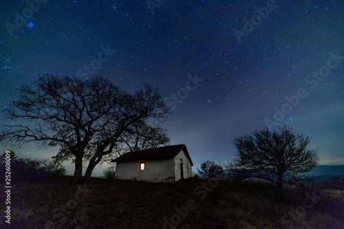 A small old church with a lighted window at night. Behind the church is an old tree without leaves. Starry Sky with Milky Way.