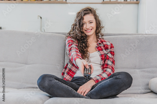 Free Download A beautiful woman watches the TV and is sitting on the couch and holds the remote control in his hand. A brunette in a shirt looks at the TV with long hair. - GFXLoad