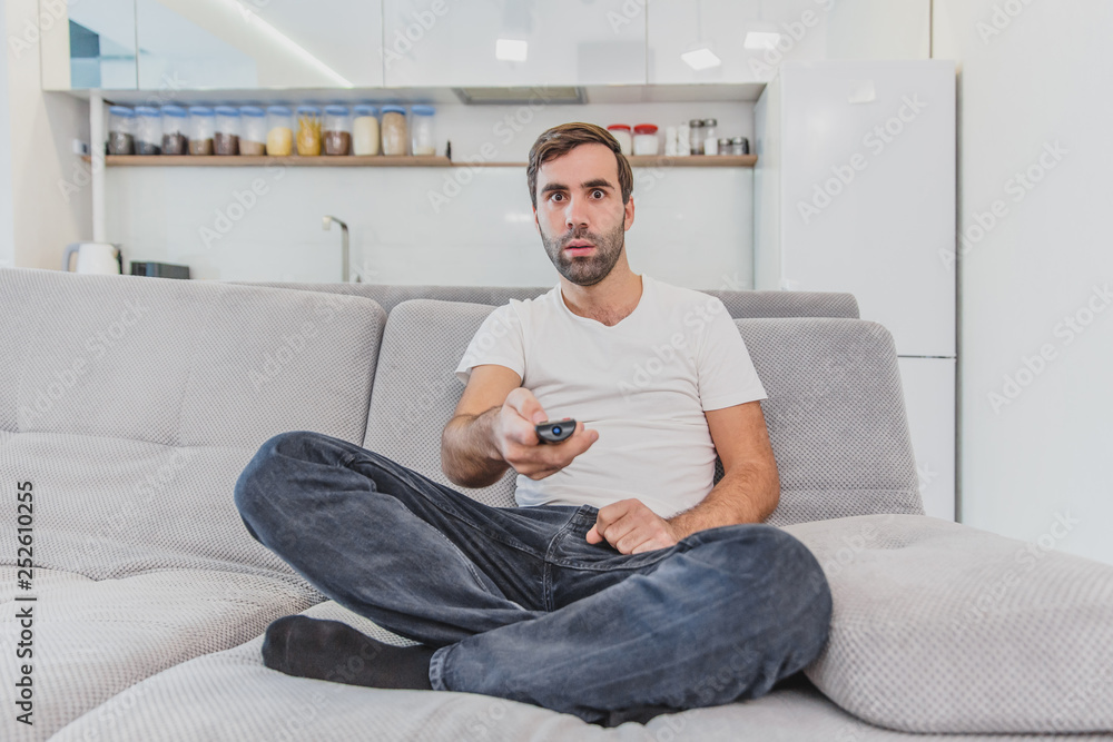 Beautiful hilarious young man holding a remote control. During this, the TV is watching while sitting on the couch at home. Beautiful hilarious young man holding a remote control. During this, the TV is watching while sitting on the couch at home.