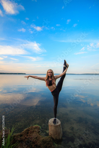 Blonde woman doing standing split outdoors on a stump in the lake. Yoga nature concept.