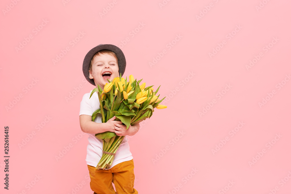 Adorable smiling child with spring flower bouquet looking at camera isolated on pink. Little toddler boy in hat holding yellow tulips as gift for mom. Copy space for text on right side