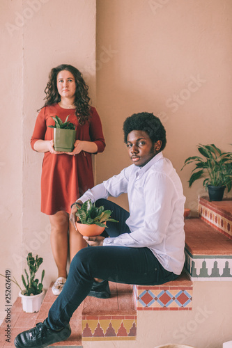 Interracial lifestyle couple portrait. Dark-skinned african man siiting on stairs with plant in pot at hands. Adult woman with home flower in hadns standing behind black nigerian male. Loving family. photo