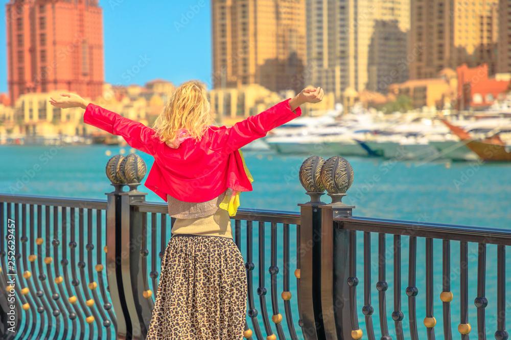 Tourism in Qatar. Blonde elegant woman at Marina walkway looks at Porto Arabia, The Pearl-Qatar's main harbor. Carefree tourist with open arms in Doha, Persian Gulf, Middle East in Arabian Peninsula.