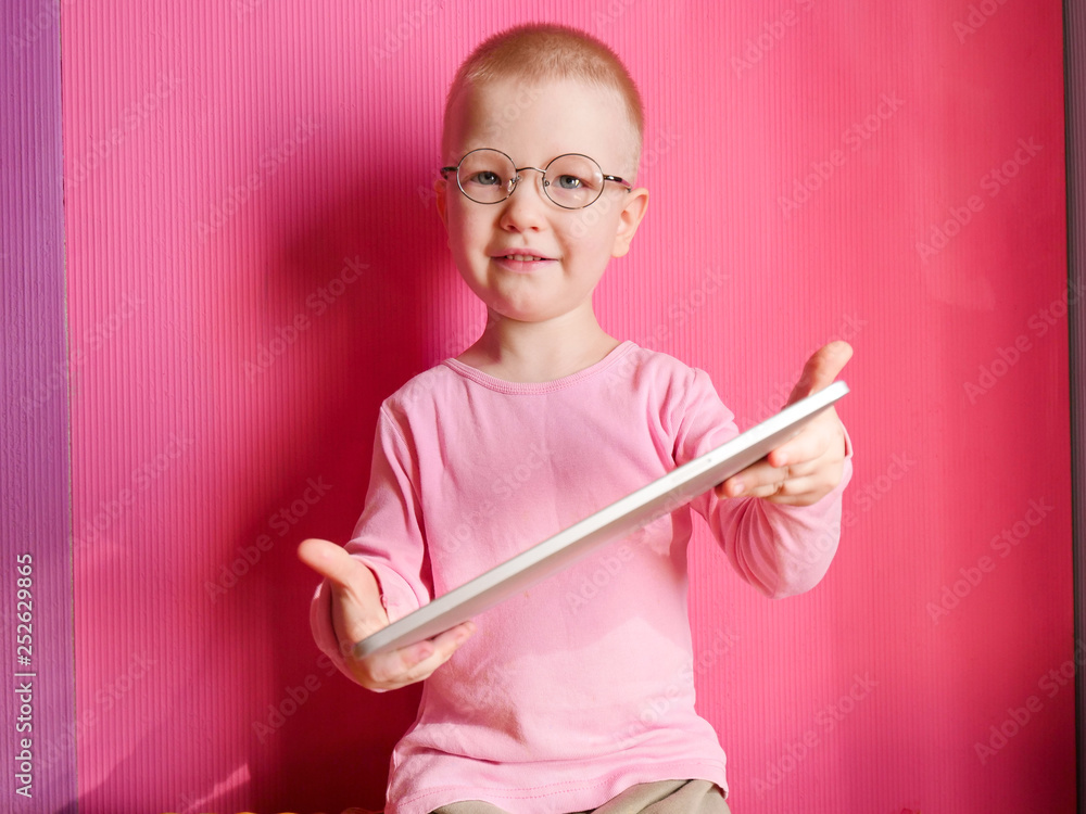 Portrait of funny child in a pink T-shirt in new glasses is playing with computer tablet on pink background. boy is proud of his glasses, clothes and shoots Portrait of funny child in a pink T-shirt in new glasses is playing with computer tablet on pink background. boy is proud of his glasses, clothes and shoots