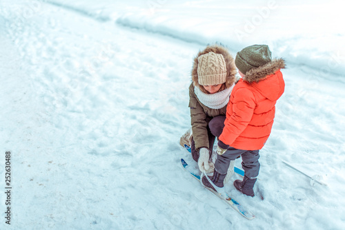 A young mother straightens her skiing attachment to her young son for 3-5 years, in the winter in city against backdrop of snow and snow drifts. Free space for text. The first steps to ski child.