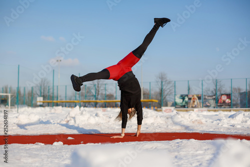Young caucasian female blonde in violet leggings stretching exercise on a red running track in a snowy stadium. fit and sports lifestyle