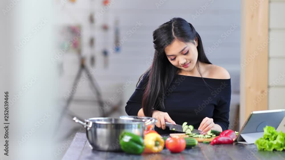 Medium shot pretty smiling young Asian woman cutting cucumber sitting at table in kitchen Medium shot pretty smiling young Asian woman cutting cucumber sitting at table in kitchen