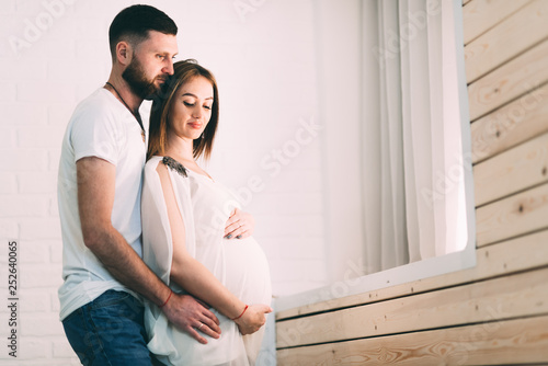 Young pregnant couple near the window of the house, on the background of a brick white wall and a wooden window sill