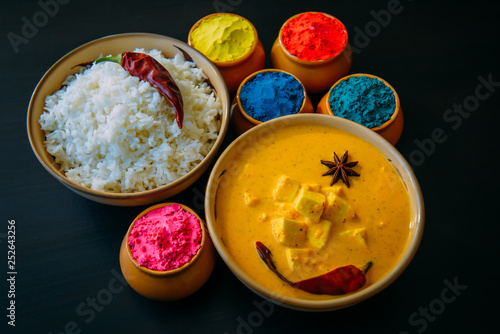 Holi indian festival of colors. Food with colours, steam rice, panir butter masala, pepper chilie, starnise. Powder colours arranged over black background. Selective focus