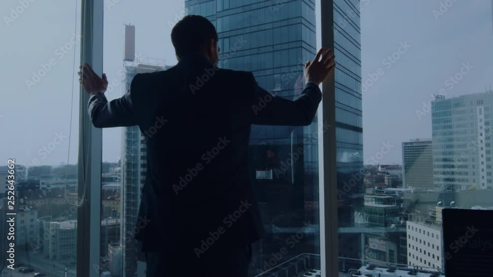 Back View of the Thoughtful Businessman wearing a Suit Standing in His Office, Contemplating Next Big Business Deal, Looking out of the Window. Big City Business District Panoramic Window View Back View of the Thoughtful Businessman wearing a Suit Standing in His Office, Contemplating Next Big Business Deal, Looking out of the Window. Big City Business District Panoramic Window View