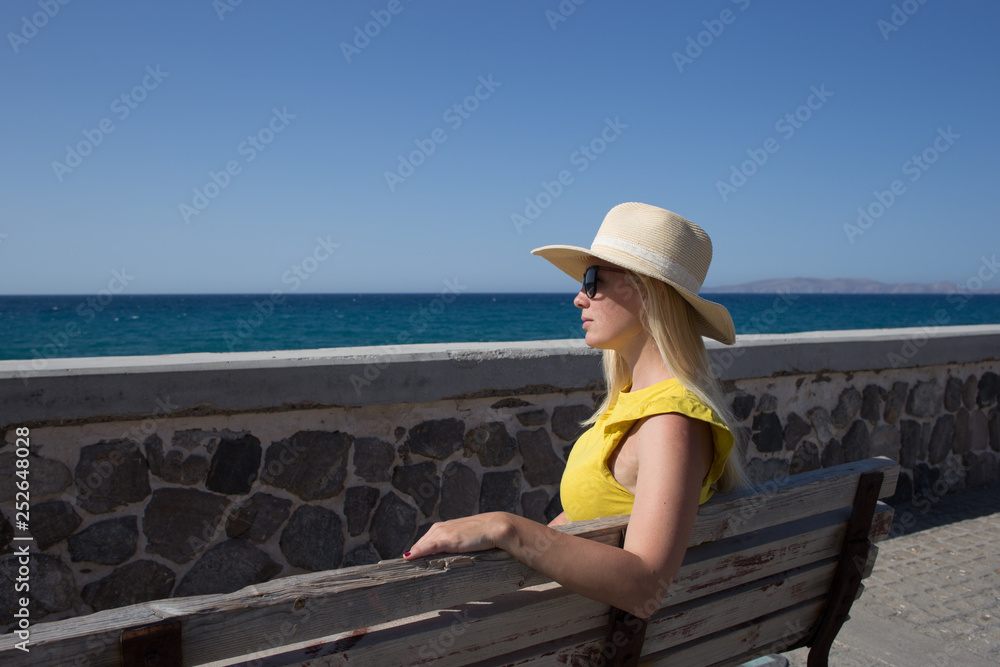 Young woman yellow t-shirt and hat resting on the seafront on a wooden bench