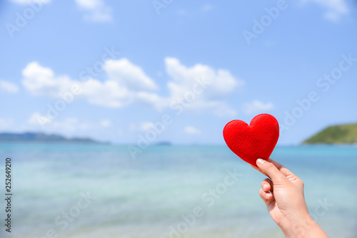 Woman hand holding a red heart on the beach with blurred sea and blue sky background. Summer day concept. Vacation holidays concept. Love concept.