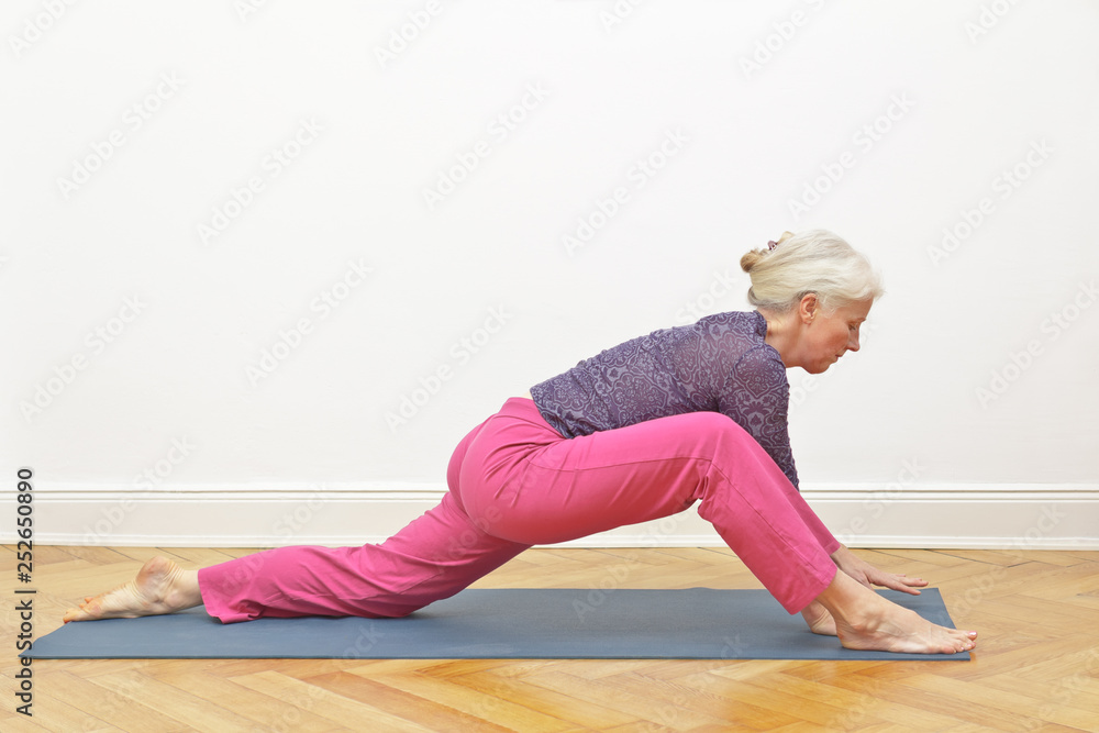 Flexible senior woman doing yoga exercise in front of a white wall, self-improvement concept, position dragon intro