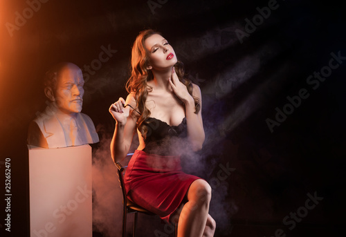 Фотография Beautiful sexy leggy busty blonde girl wearing a red skirt and black body sits on a chair at the statue of the communist leader Lenin in theatrical smoke