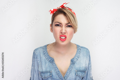 Portrait of dissatisfied beautiful young woman in casual blue denim shirt with makeup and red headband standing and looking at camera clenching teeth. indoor studio shot, isolated on white background.