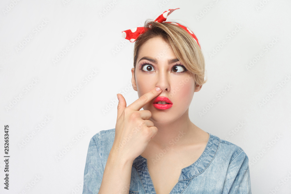 Closeup portrait of funny crazy crossed eyes young woman in casual blue denim shirt with makeup and red headband standing looking and picking her nose. indoor studio shot, isolated on white background Closeup portrait of funny crazy crossed eyes young woman in casual blue denim shirt with makeup and red headband standing looking and picking her nose. indoor studio shot, isolated on white background