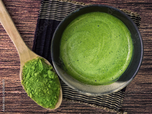 A cup of green tea latte on bamboo mat and matcha powder in spoon on wooden background.