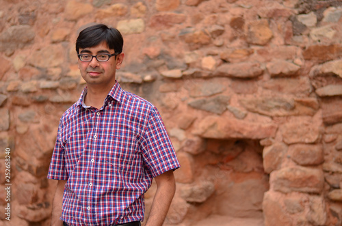 Portrait of a confident looking young Indian man wearing spectacles standing against a brown rock wall in Delhi India