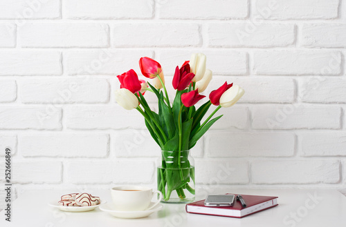 tulip flowers are in a vase on the table, cup of coffee, diary and smartphone, white brick wall as background
