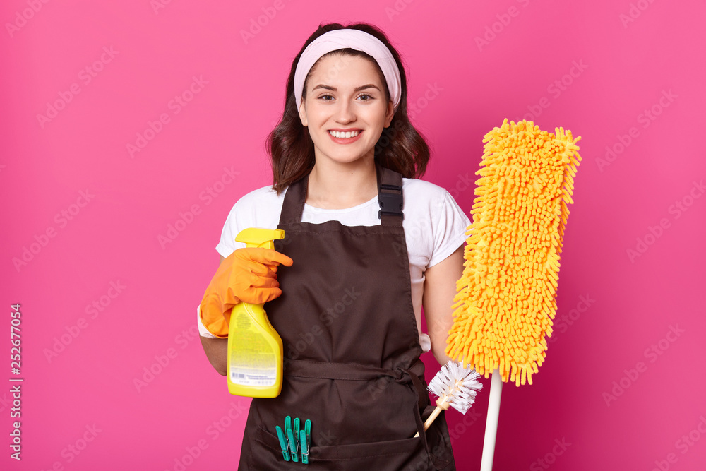 Cheerful young housewife wears white t shirt, brown apron with green clothespins, orange rubber gloves for hands protection. Holds spray and mop. Smiling female cleans house. Works at home. Cheerful young housewife wears white t shirt, brown apron with green clothespins, orange rubber gloves for hands protection. Holds spray and mop. Smiling female cleans house. Works at home.