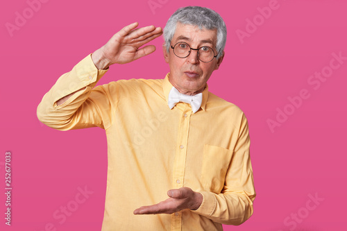 Calm senior shows large or big object, stands against pink wall, dressed in stylish yellow shirt and white bowltie, has serious facial expression. Pensioner shapes square. Male demonstrates size. photo