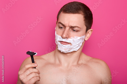 Indignant handsome young European man looks at sharp razor, has white foam gel on face, morning routine in bathroom, poses with nude body, isolated over pink background. Facial treatment concept photo