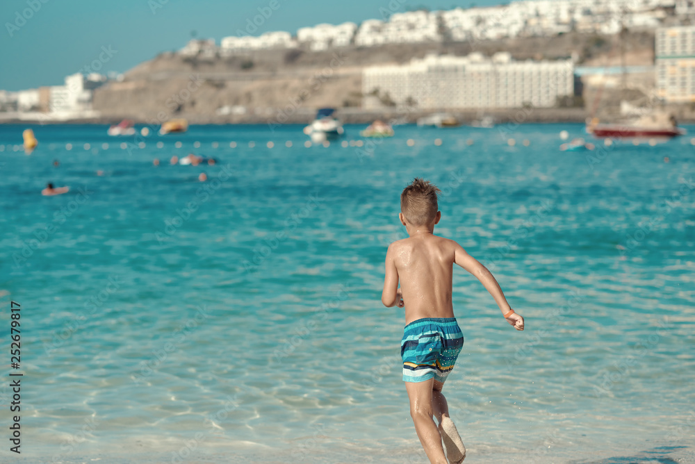 Active European boy in a striped swimming shorts is running towards the sea. He is having fun spending his holidays on the seashore.