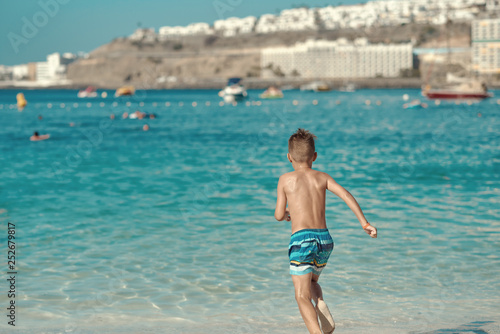 Active European boy in a striped swimming shorts is running towards the sea. He is having fun spending his holidays on the seashore.