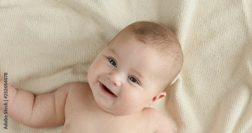Smiling little baby boy with big brown eyes lying on the bed, looking at camera and smiling. 4k view from above Smiling little baby boy with big brown eyes lying on the bed, looking at camera and smiling. 4k view from above