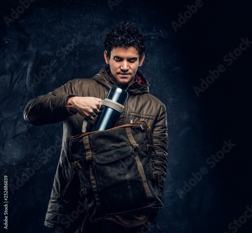 Young traveler in hiking gear puts a thermos in his backpack in the studio against a dark wall  © Fxquadro