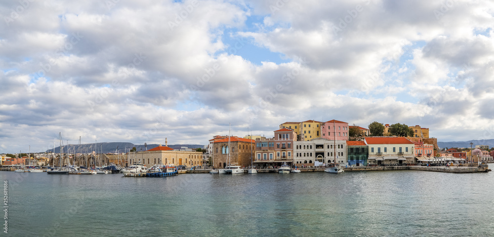 A panorama of the seaport town Chania, the island of Crete, Greece. A harbor with wooden pantons, moored yachts, ships, boats. Colorful architecture of modern and old houses. Mountains on the horizon.