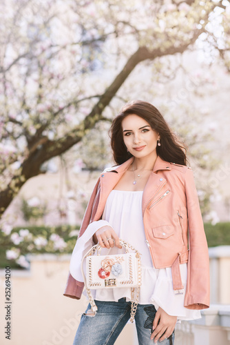 Outdoor portrait of young beautiful fashionable lady wearing pink leather jacket, holding small stylish white bag with floral application. Blooming tree on background. Spring fashion concept