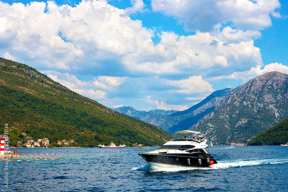 Incredible bright seascape. View of green wooded mountains and blue sea, blue sky and white clouds and a yacht sailing through the waves between the mountains. Boka Kotorska Bay, Montenegro