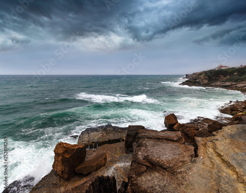 Landscape of the Atlantic Ocean before the storm. Big waves crashing into the rocky shore . Dramatic view of the dark overcast sky. Westernmost extent of Europe. Portugal. 
