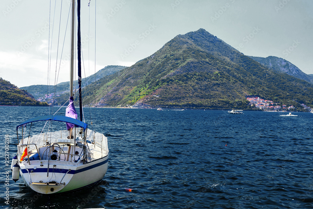 Incredible bright seascape. View of green wooded mountains and blue sea, blue sky and white clouds and a yacht sailing through the waves between the mountains. Boka Kotorska Bay, Montenegro