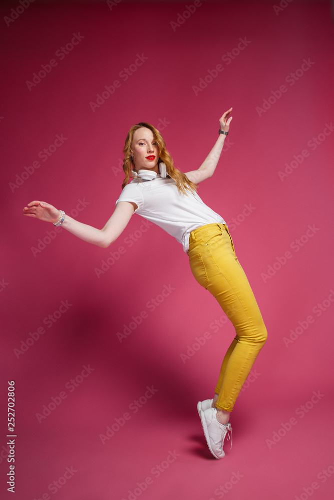 Obraz premium Good Party Mockup. Cheerful blonde woman in white headphones, white T-shirt and trendy yellow jeans dancing on pink background.