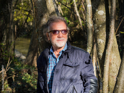Senior man with sunglasses, beard and white mustache, posing in the middle of the forest.